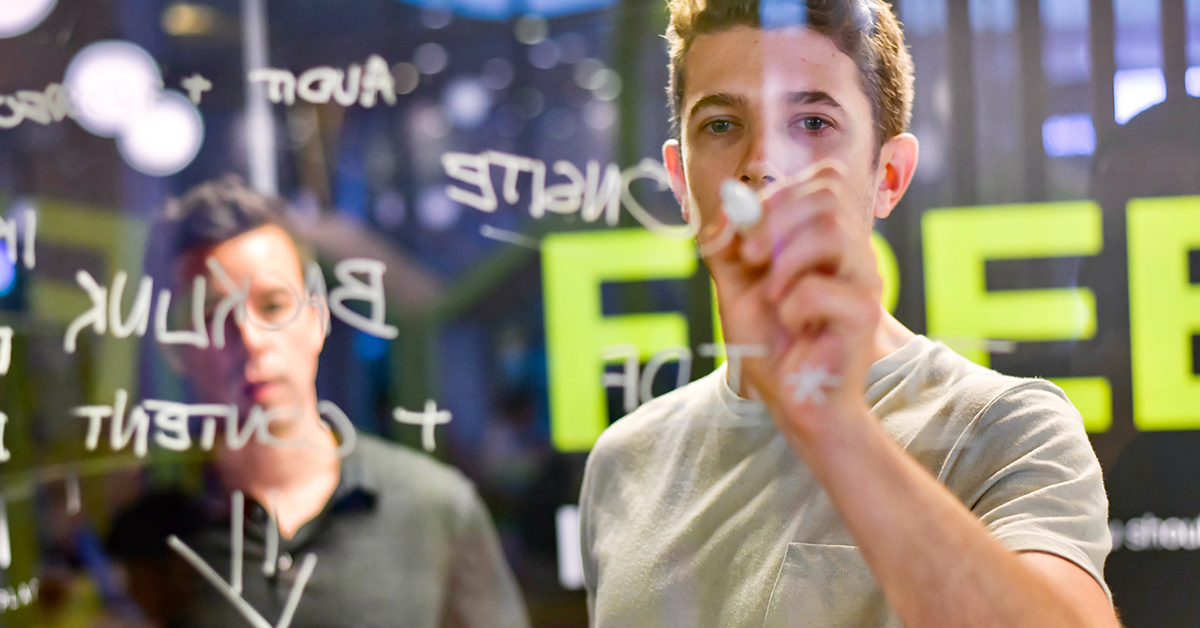 two people writing on a clear board with dry erase markers